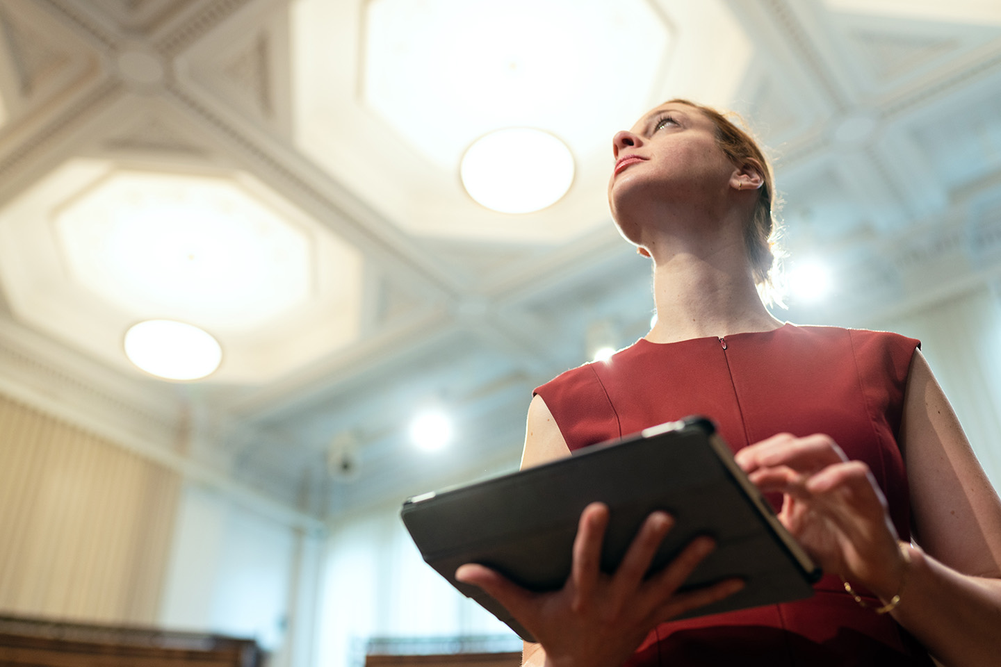 Woman with a red top on looking up at a stately ceiling holding a work tablet