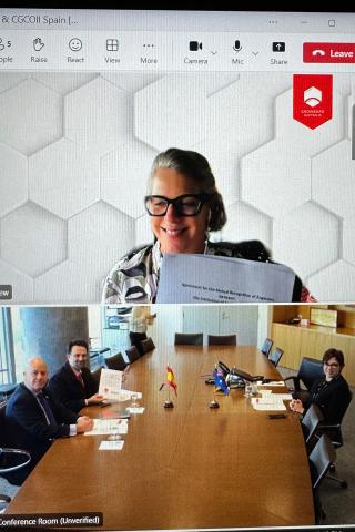 Screenshot of a video call with two participants. One box shows one woman, the other shows a board room with three people stat at a large table